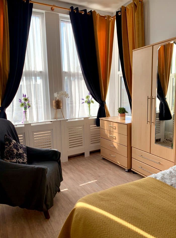 A bright bedroom with tall windows, an armchair, a chest of drawers and a wardrobe