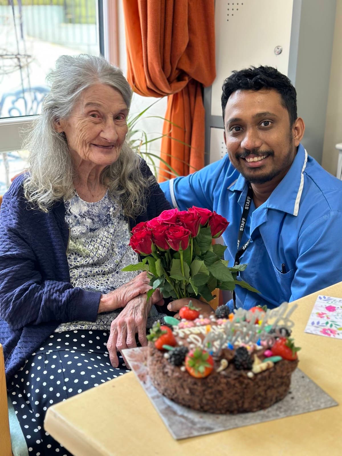 A carer sitting with a resident beside a bouquet of red roses and a cake